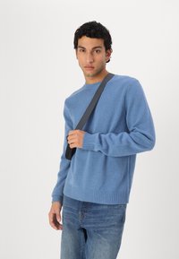 Man i blå tröja och jeans står och håller det svarta bandet till en axelväska mot en vit bakgrund.