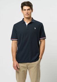 Laivastonsininen lyhythihainen polo-paita, valkoinen kaulus, punavalkoraidalliset hihansuut, brodeerattu logo rinnassa, teksturoitu kangas, yhdistetty vaaleisiin housuihin.