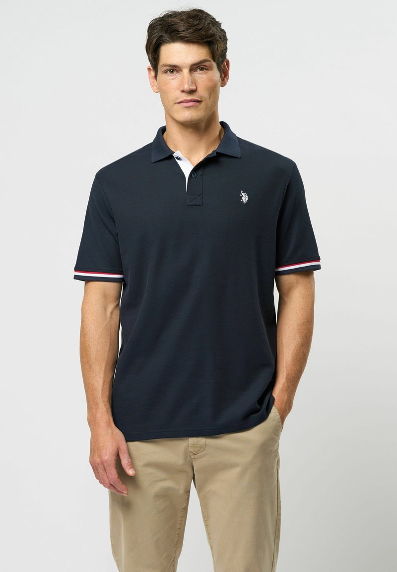 Laivastonsininen lyhythihainen polo-paita, valkoinen kaulus, punavalkoraidalliset hihansuut, brodeerattu logo rinnassa, teksturoitu kangas, yhdistetty vaaleisiin housuihin.