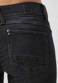 Dark denim jeans feature a back pocket with two metal rivets, black leather logo patch at the waistband, and subtle stitching details.