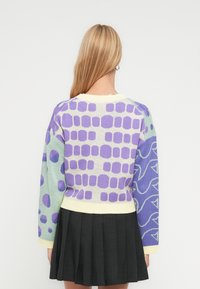 Ξανθιά γυναίκα με πλάτη, φοράει πολύχρωμο πουλόβερ με μοτίβα σε μωβ και πράσινες αποχρώσεις και μαύρη πλισέ φούστα.