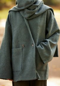 Zelený, strukturovaný vlnený kabát s velkou kapucí, oversize střih, dvě přední knoflíky a kapsa na levé straně.