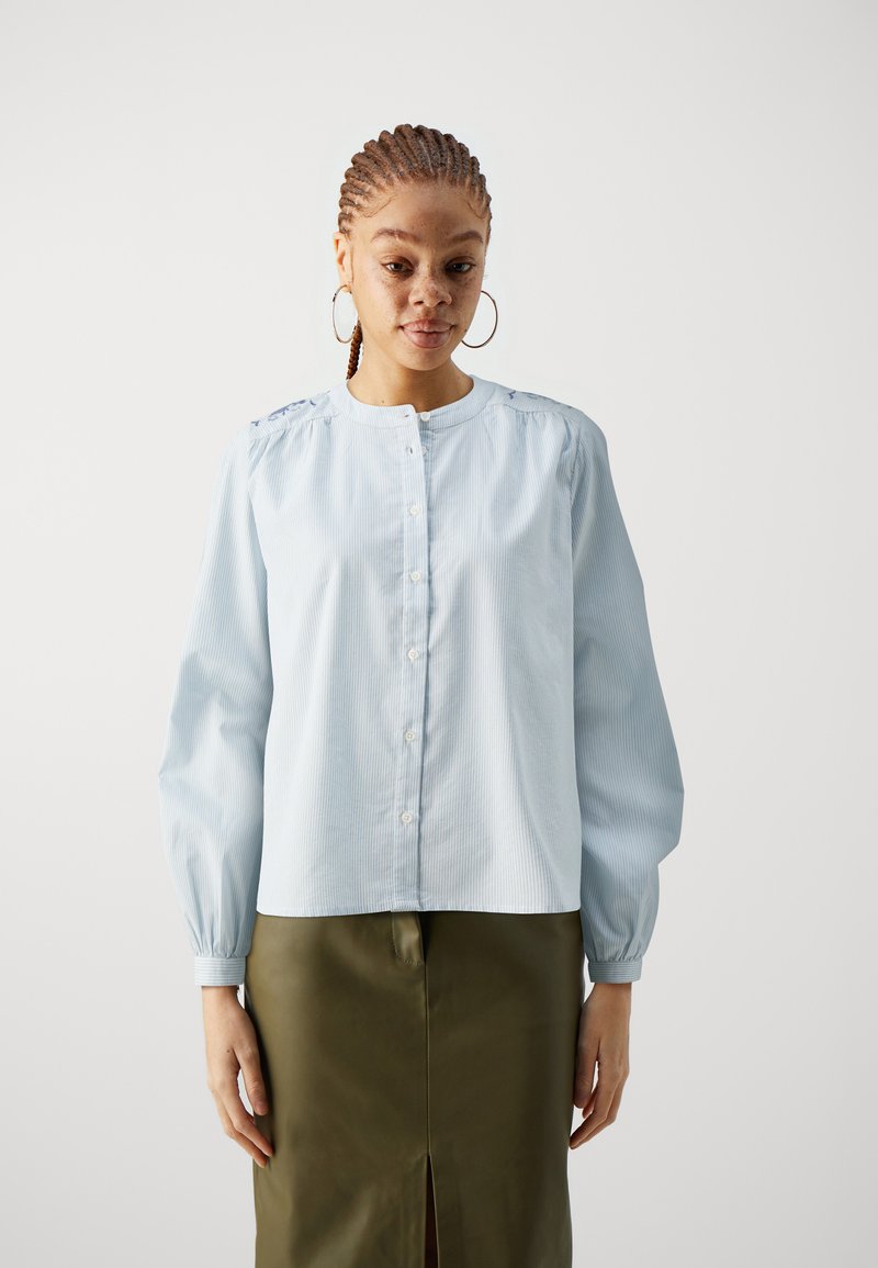 Camicia azzurra a righe con bottoni, maniche lunghe, scollatura rotonda e dettaglio arricciato sulle spalle, abbinata a una gonna verde oliva.