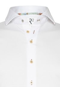 Witte polo met een klassieke kraag, voorzien van beige knopen en een bloemenpatroon aan de binnenkant van de kraag. Soepele textuur, casual ontwerp.