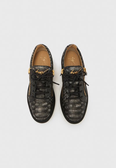 Svarta sneakers i texturerat läder med guldfärgade sidodragkedjor, svarta snören och en guldfärgad logotyp på tungspetsen, visade uppifrån på en vit bakgrund.