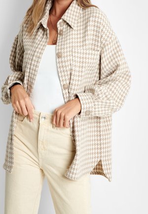 Kobieta w beżowo-białej kurtce koszulowej w kratę piórkową na białej bluzce i kremowych spodniach, poprawiająca pasek spodni.