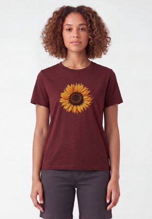 Femme aux cheveux bouclés portant un t-shirt bordeaux avec un grand motif tournesol jaune, associé à un pantalon gris foncé, debout devant un fond blanc.
