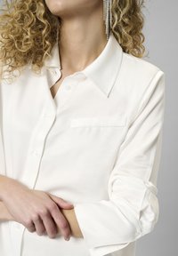 Eine lockige Person trägt ein weißes Hemd mit gekrempelten Ärmeln und langen, herabhängenden Strass-Ohrringen und hält einen Arm.
