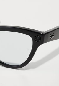 Occhiali da sole neri con forma rettangolare, lenti trasparenti e una fotocamera incorporata sul lato destro. Presentano il logo Ray-Ban sulla stanghetta.