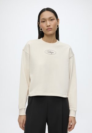 Femme aux cheveux noirs raides portant des boucles d'oreilles créoles dorées, un sweat crème avec le logo "Hugo" et un pantalon noir sur un fond uni.