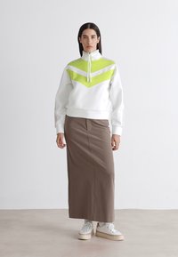 Fehér pulóver zöld részletekkel, cipzáras nyakkal és chevron mintázattal; barna midi szoknyával és fehér sportcipővel párosítva. Sima textúra.