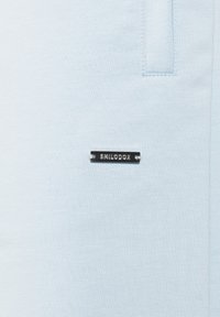 Tejido azul claro con una textura suave, que presenta una placa de logo rectangular negra con la inscripción "SMILODOX". Diseño minimalista sin patrones adicionales.