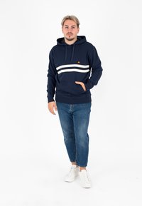 Felpa navy con righe orizzontali bianche, tasca a marsupio e cordino. Abbinata a jeans blu e sneaker bianche. Il modello si trova su uno sfondo bianco.
