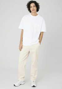 T-shirt blanc en coton avec une poche sur la poitrine, associé à un pantalon beige à côtes et des baskets gris clair. Le modèle se tient de manière décontractée avec les mains dans les poches.