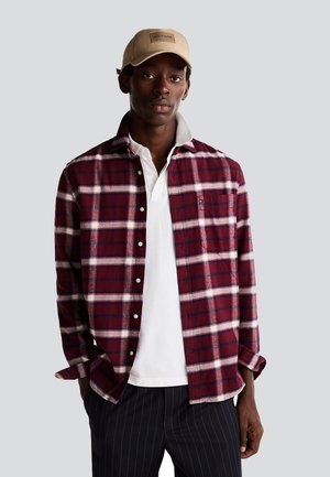 Camicia a quadri rossa e bianca con bottoni, indossata sopra una polo bianca, abbinata a pantaloni scuri a righe e un berretto beige.