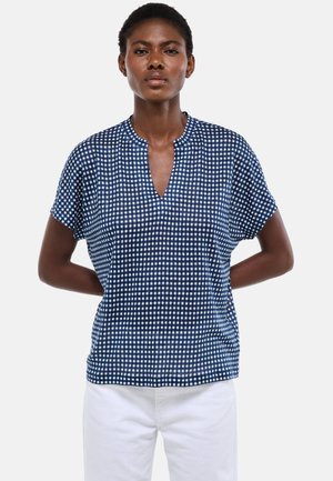 Femme portant un chemisier à manches courtes à carreaux bleu marine et blanc avec un col fendu, associé à un pantalon blanc, debout devant un fond uni.