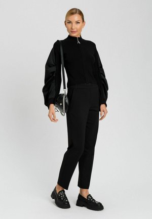 Maglione nero con zip e maniche a sbuffo, pantaloni neri su misura e scarpe nere massicce. Piccola borsa nera con tracolla in catena argentata.