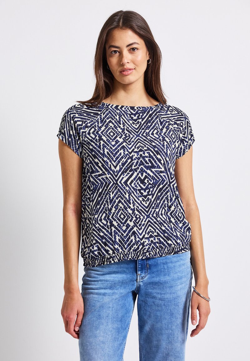 Top de manga corta con un patrón abstracto en azul marino y blanco, escote redondo, dobladillo elástico, combinado con jeans de color azul claro. Tejido suave y ligero.