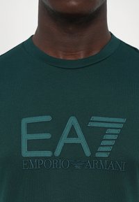Tumši zaļš T-krekls ar izšūtu EA7 Emporio Armani logotipu uz krūtīm, ko valkā cilvēks ar tumšu ādu, tuvplānā uz apkakles un augšējā ķermeņa.