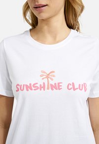 Woman wearing white t-shirt with pink text "SUNSHINE CLUB" and a small embroidered palm tree replacing the letter "I".