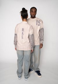 Béžová košeľa s dlhým rukávom so sivými akcentmi; obsahuje text "M.A.S." a "15." Dopĺňa ju svetlomodré rifle; voľný strih, ležérny dizajn.