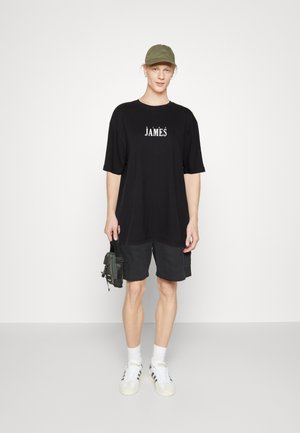 Camiseta negra de gran tamaño con estampado de "JÅMËS", combinada con pantalones cortos oscuros. Lleva una gorra olivo y zapatillas blancas, sosteniendo un pequeño bolso negro.
