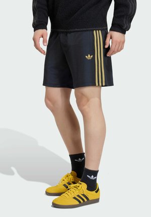 Osoba ubrana w czarne szorty ze złotymi paskami Adidas, czarne skarpetki Adidas i żółte buty sportowe Adidas, stojąca na jasnym tle.