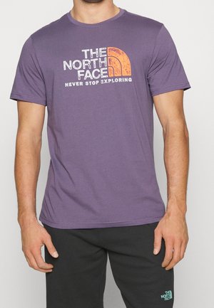 Mann trägt ein lila kurzärmeliges T-Shirt mit "The North Face Never Stop Exploring"-Logo und schwarze Hose mit kleinem Logo.