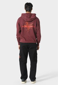 Felpa bordeaux con grafica arancione "PAS DE MONACO" sulla schiena, abbinata a pantaloni cargo neri con tasche laterali e dettagli del logo.