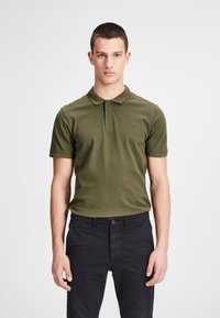 Olivgrünes Poloshirt mit kurzen Ärmeln und Kragen, ausgestattet mit zwei Knöpfen am Hals, Markenlogo auf der Brust, kombiniert mit dunklen Hosen.