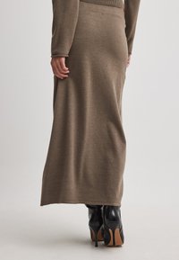 Jupe maxi en tricot marron avec une taille ajustée et une fente sur le côté, associée à des bottes à talons hauts noires, vue de l'arrière.