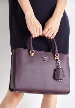 Hände halten eine lilafarbene Guess-Handtasche mit goldfarbenen Beschlägen, getragen von einer Person in einem dunklen Kleid vor einem schlichten hellen Hintergrund.