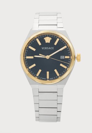 Sølvfarvet metalarmbåndsur med sort urskive, guldfarvet ramme, guldfarvede timemarkeringer, datovisning klokken tre og "Versace"-logo under tolv.