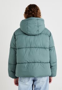 Svijetlozeleni puffer jakna s kapuljačom, sa horizontalnim kvadratnim sekcijama i mekanom, glatkom teksturom. Nosi se preko plavih traperica.