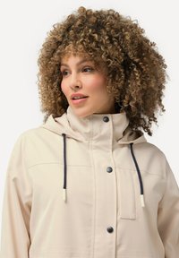 Giacca impermeabile beige con cappuccio, dotata di lacci neri e dettagli con bottoni. Tessuto liscio con tasca frontale.