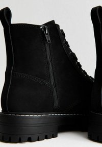 Bottines en daim noir avec fermeture éclair sur le côté, lacets à l'avant, semelle en caoutchouc épaisse avec motifs et détail de couture blanche.