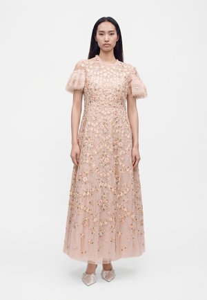 Femme aux longs cheveux foncés portant une robe longue en dentelle rose à motifs floraux avec des manches courtes bouffantes et des chaussures en satin pointues, debout sur un fond blanc.