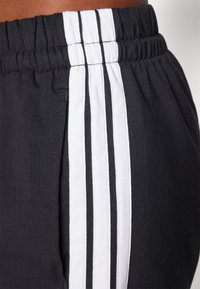 Gros plan sur un pantalon de sport noir avec trois rayures blanches et une taille élastique au niveau de la taille et des hanches.