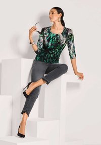 Schwarz-grün gemusterte Bluse mit Dreiviertelärmeln, kombiniert mit dunkelgrauen Jeans und schwarzen High Heels, sitzt auf weißen geometrischen Stufen.