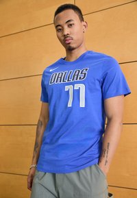 Blå Nike t-skjorte med "DALLAS" og "77" i hvit skrift. Korte ermer, rund hals, glatt tekstur. Modell iført olivengrønne shorts.