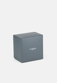 Boîte de montre Timex, forme carrée, matériau en carton gris foncé, texture lisse, avec le mot "TIMEX" imprimé en blanc sur le devant.
