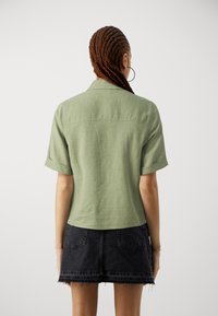Camisa de lino verde de manga corta con cuello de solapa, vista desde atrás, combinada con una falda vaquera negra de talle alto.