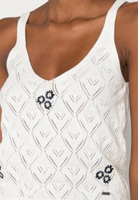 Vitt stickat linne med ett hjärtformat mönster, prytt med marinblå broderade blommor som accenter och tunna axelremmar.