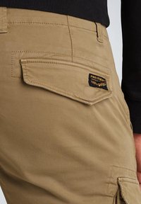 Close-up van kaki broek met een klepzak en PME Legend-logo patch op de achterkant, gedragen met een zwarte top.