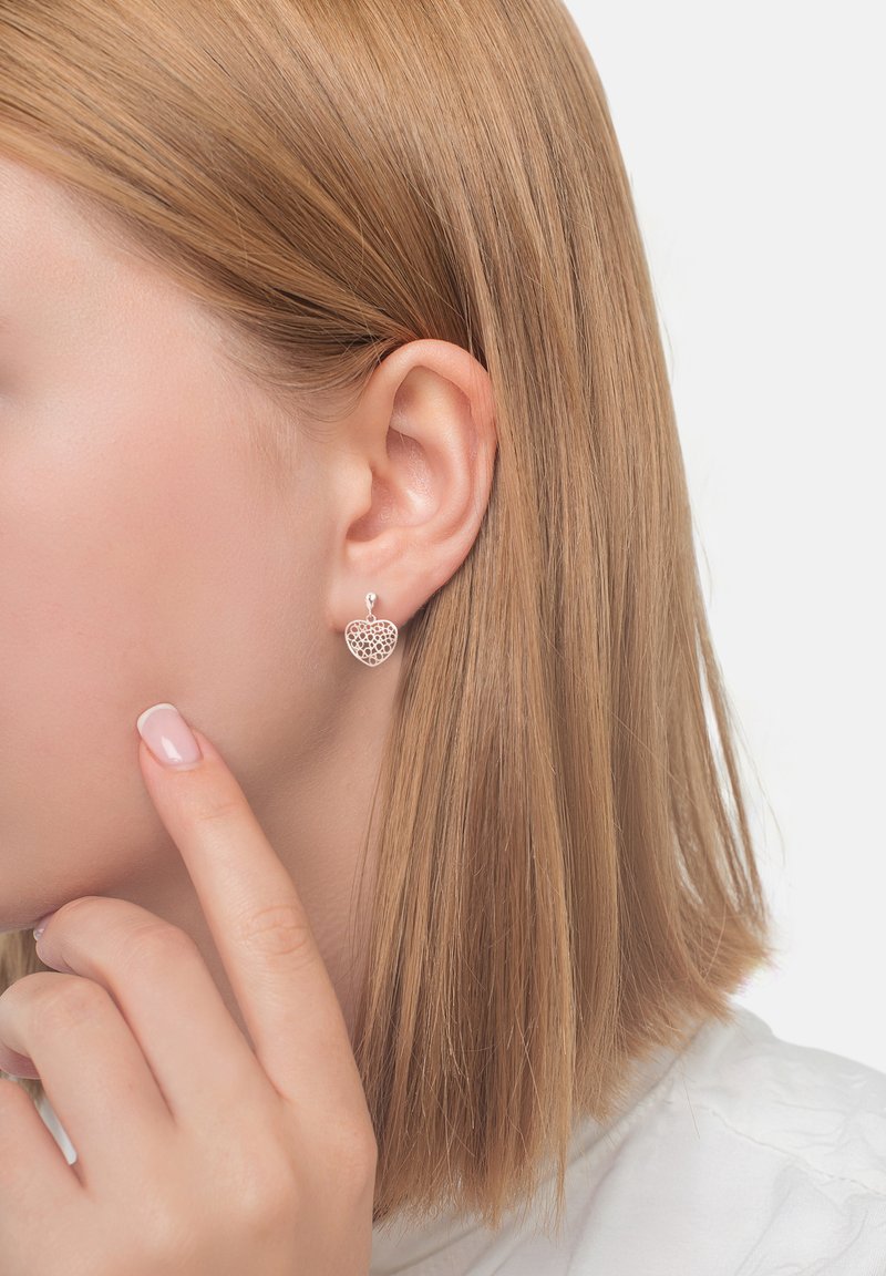 Silverfärgade örhängen i form av hjärtan med ett intrikat mönster av utskärningar, hängande från en liten ögla. Visas på en modell med rakt blont hår.