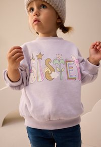Gaiši violeta sporta džemperis ar krāsainu, rotaļīgu "SISTER" grafiku, kurā attēlots vienradzis, varavīksne un ziedu motīvi. Ar ribām apdarinātas aproces un apakšmala.