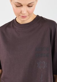 Mørk lilla kortærmet t-shirt lavet af blød stof, med et grafisk print og tekst på venstre side, og en rund halsudskæring.
