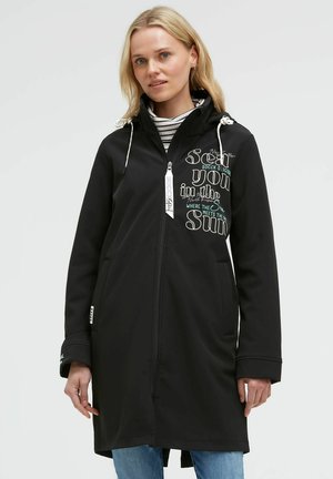 Woman wearing a black hooded long jacket with text on chest, zipped front, white drawstrings, and light blue jeans.