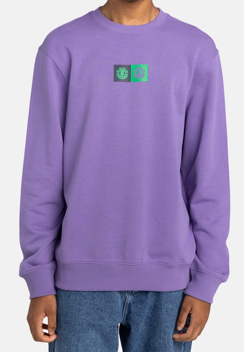 Homme portant un sweat-shirt à manches longues violet avec un petit logo carré noir et vert sur la poitrine, assorti à un jean bleu.