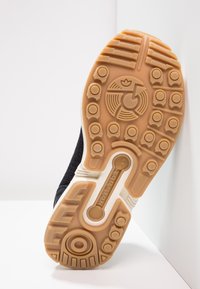 Zwarte sneakerzool met een gestructureerd rubber en een gepatternde grip, met een beige tussenzool en een rond logo in het midden.